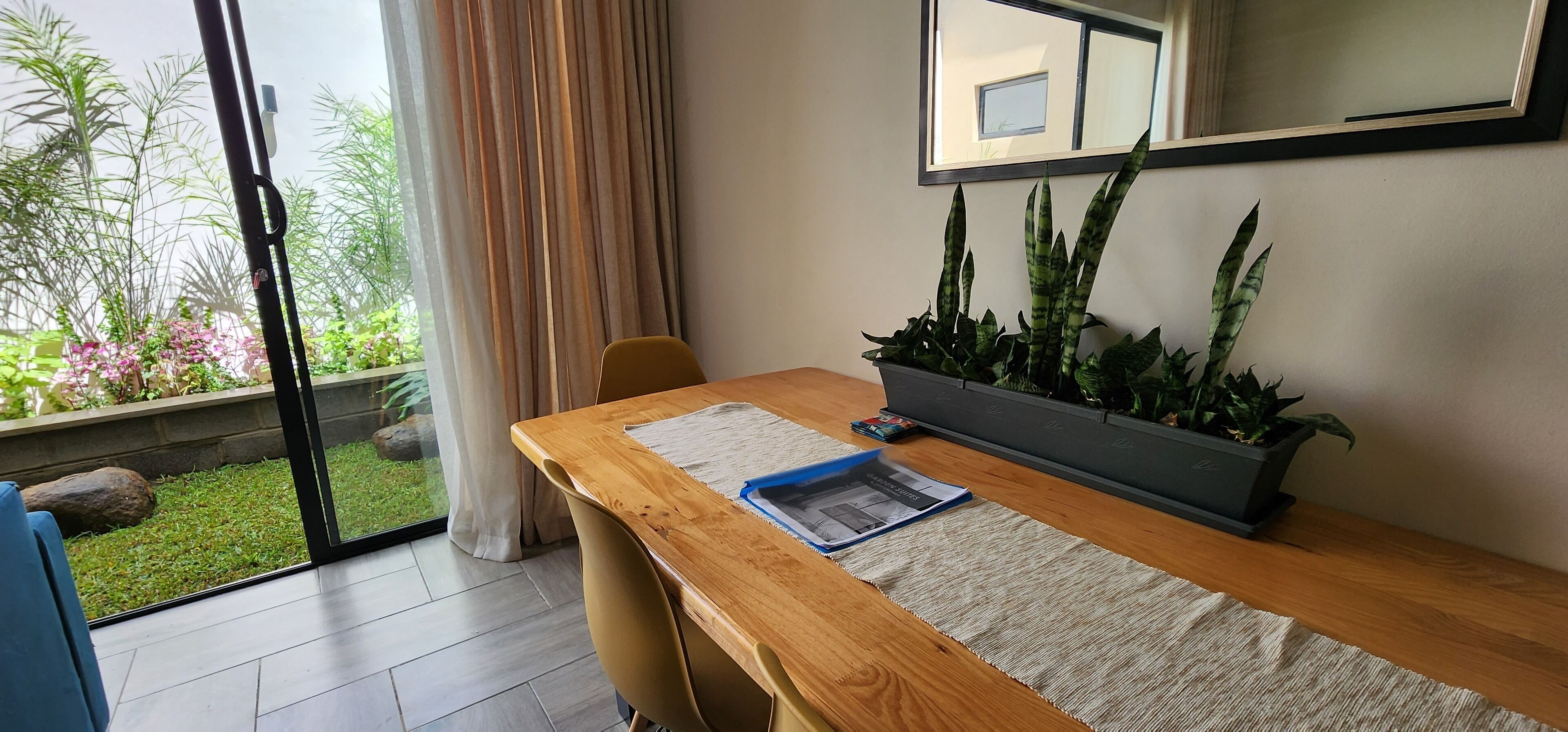 Une pièce avec un bureau en bois, une plante en pot, une chaise, une fenêtre avec des rideaux et une vue sur la verdure à l’extérieur.