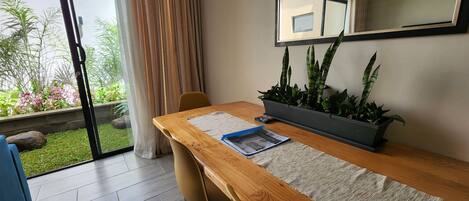 Une pièce avec un bureau en bois, une plante en pot, une chaise, une fenêtre avec des rideaux et une vue sur la verdure à l’extérieur.