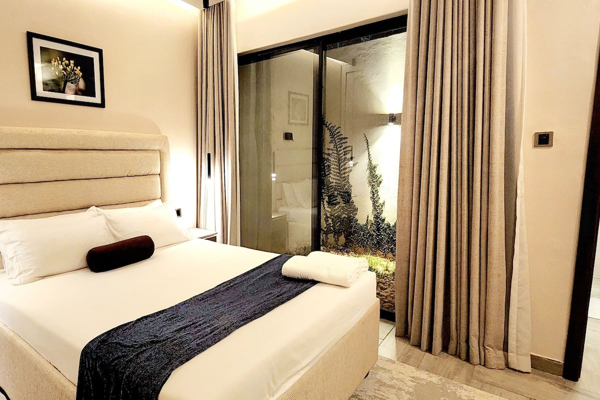 Une chambre d’hôtel moderne avec un grand lit, une petite table de chevet, un plaid sombre et une vue sur un jardin, visible à travers une porte vitrée.