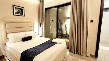 A modern hotel room with a large bed, a small bedside table, a dark throw blanket, and a view of a garden through a glass door.