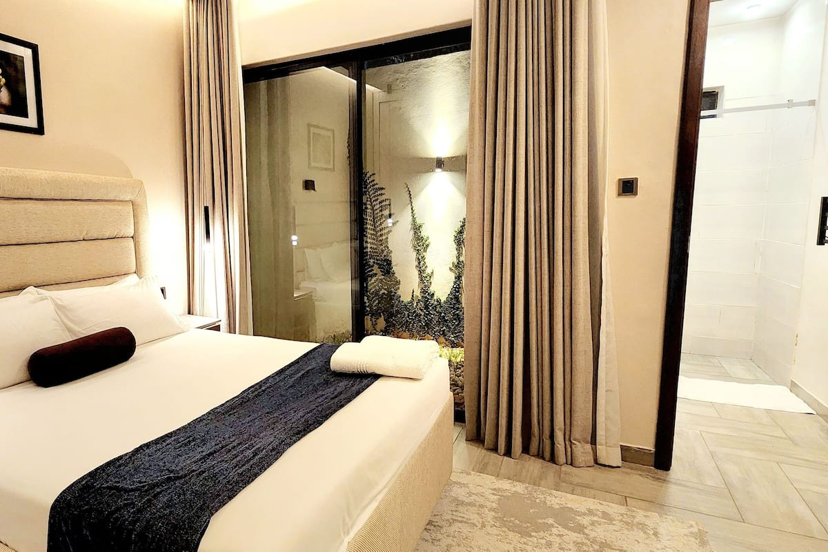 Une chambre d’hôtel avec un grand lit, une couverture épaisse de couleur sombre, une tête de lit, un tableau encadré au mur et une vue sur un jardin, visible à travers une porte vitrée.