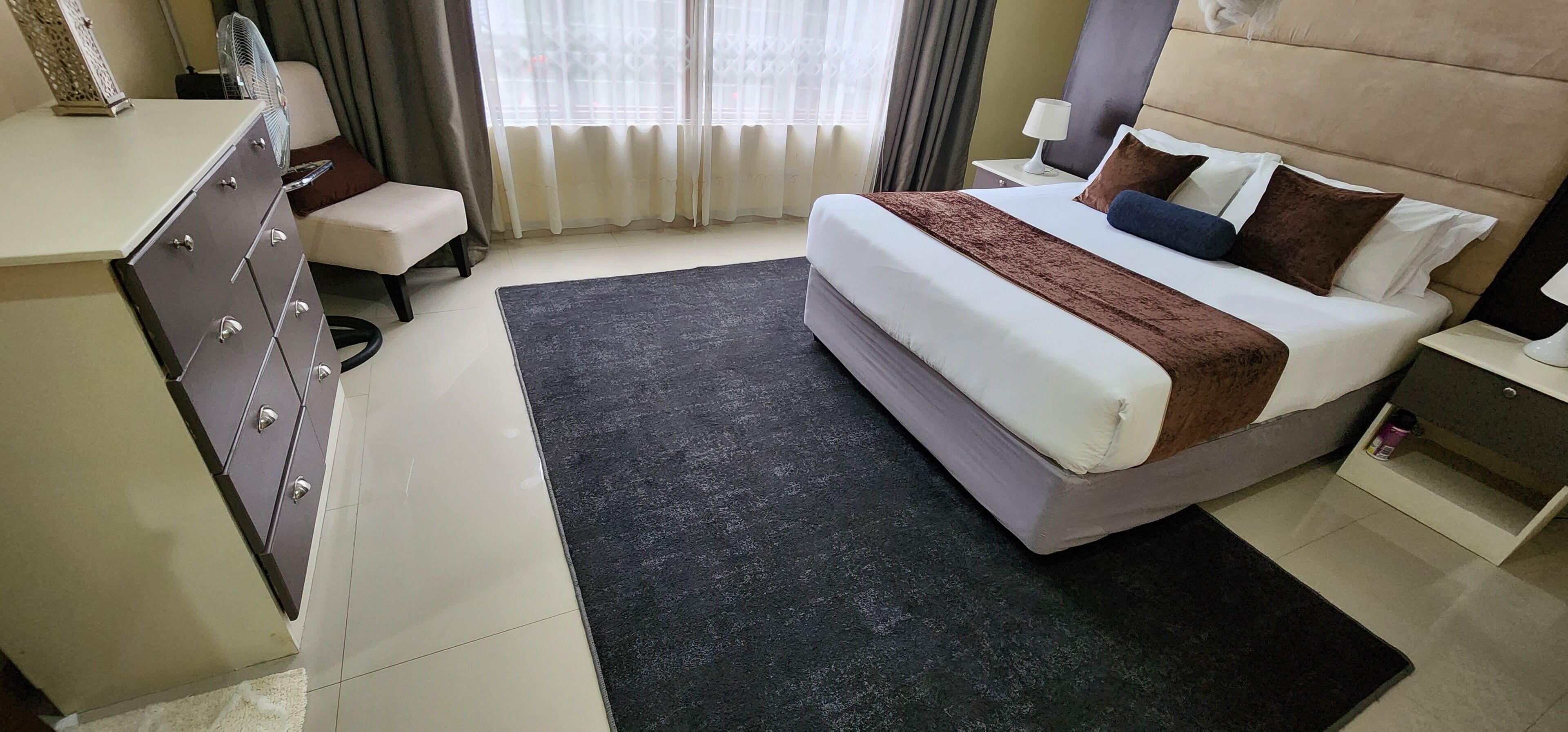 A hotel room with a bed, bedside table, lamp, and a desk with drawers.