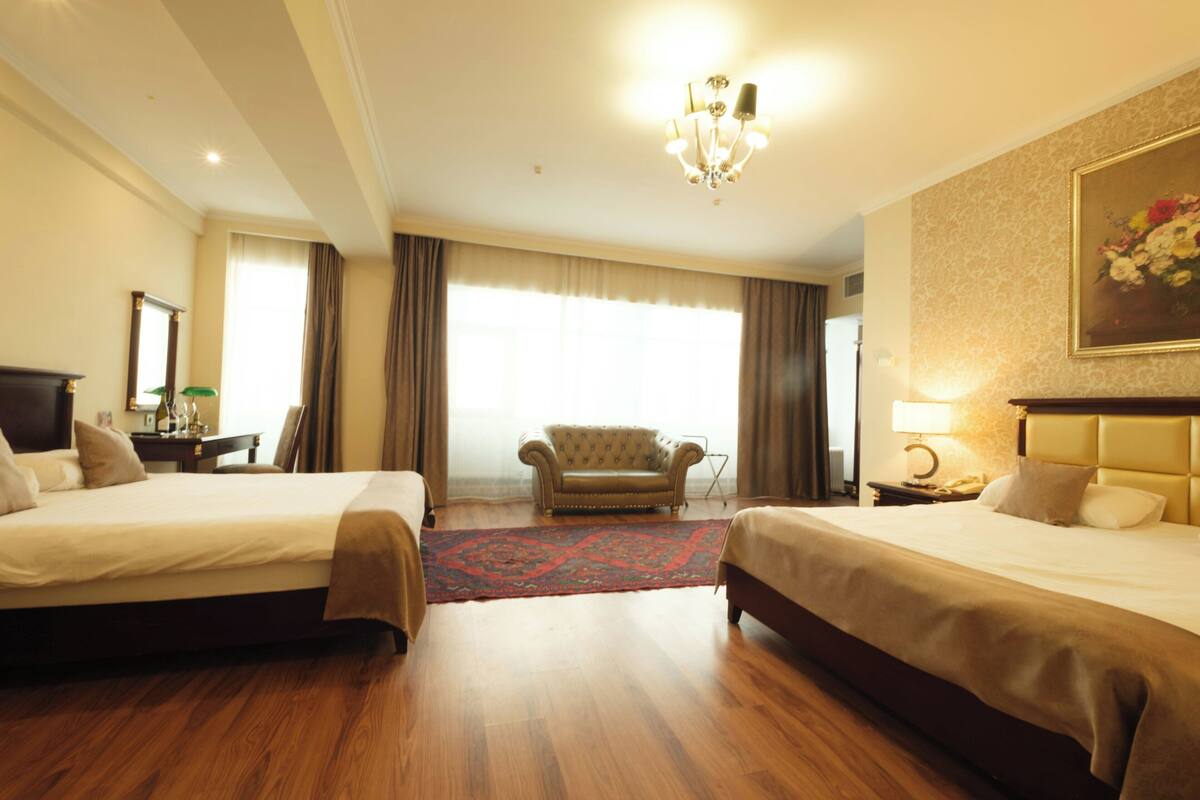 A hotel room with two beds, a chandelier, a large window with curtains, a sofa, and a painting on the wall.