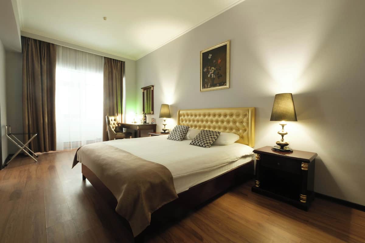 A hotel room with a large bed, two bedside lamps, a painting on the wall, and a desk with a chair.