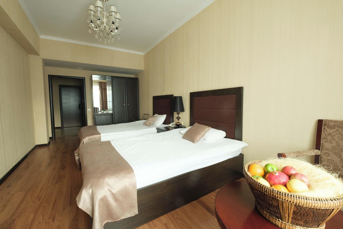 A hotel room with two beds, a chandelier, a mirror, a bedside table with a lamp, and a fruit basket.