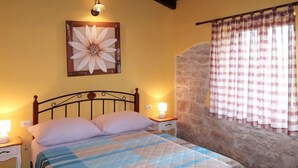 A bedroom with a bed, a nightstand, a lamp, a wall-mounted flower painting, a stone wall, and a window with checkered curtains. - Vacation home Milena by Interhome (Barban)