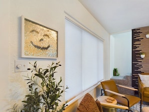 Una sala de estar moderna con un ventanal grande, una obra decorativa en la pared, una planta en maceta y una zona de estar con una silla y una mesita.