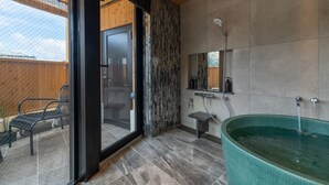 Une salle de bain moderne avec une grande baignoire, une douche et un petit lavabo. Une porte en verre donne accès à un espace extérieur agrémenté d’un banc.
