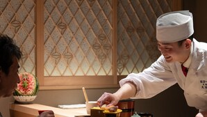 Une personne vêtue d’un uniforme blanc sert de la nourriture à une autre personne assise à une table en bois, avec un mur décoratif en arrière-plan.