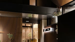 Une entrée moderne, avec une façade en bois, une enseigne ornée de caractères japonais et un petit jardin agrémenté de rochers et de plantes.