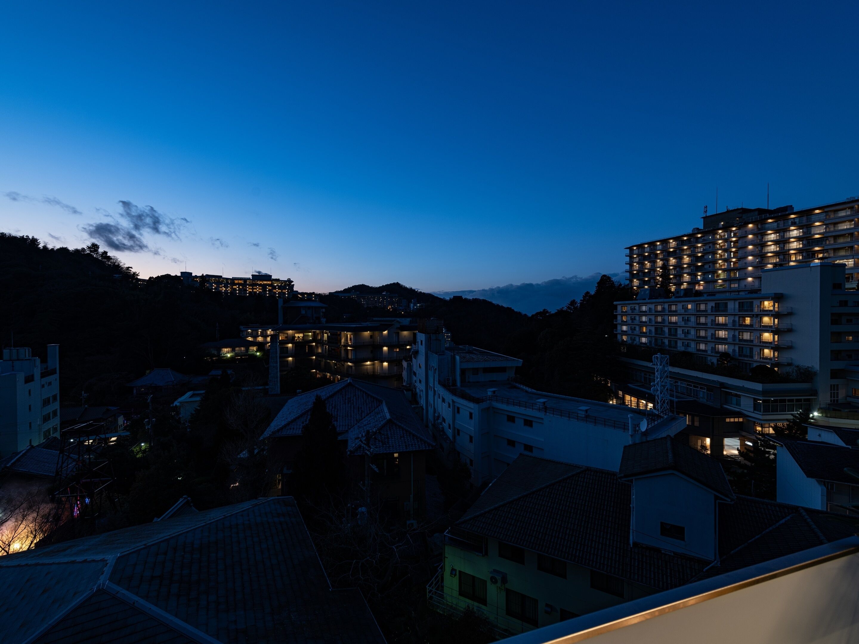 Un paisaje urbano al anochecer con varios edificios, incluyendo un hotel grande con ventanas iluminadas, rodeado de construcciones residenciales.