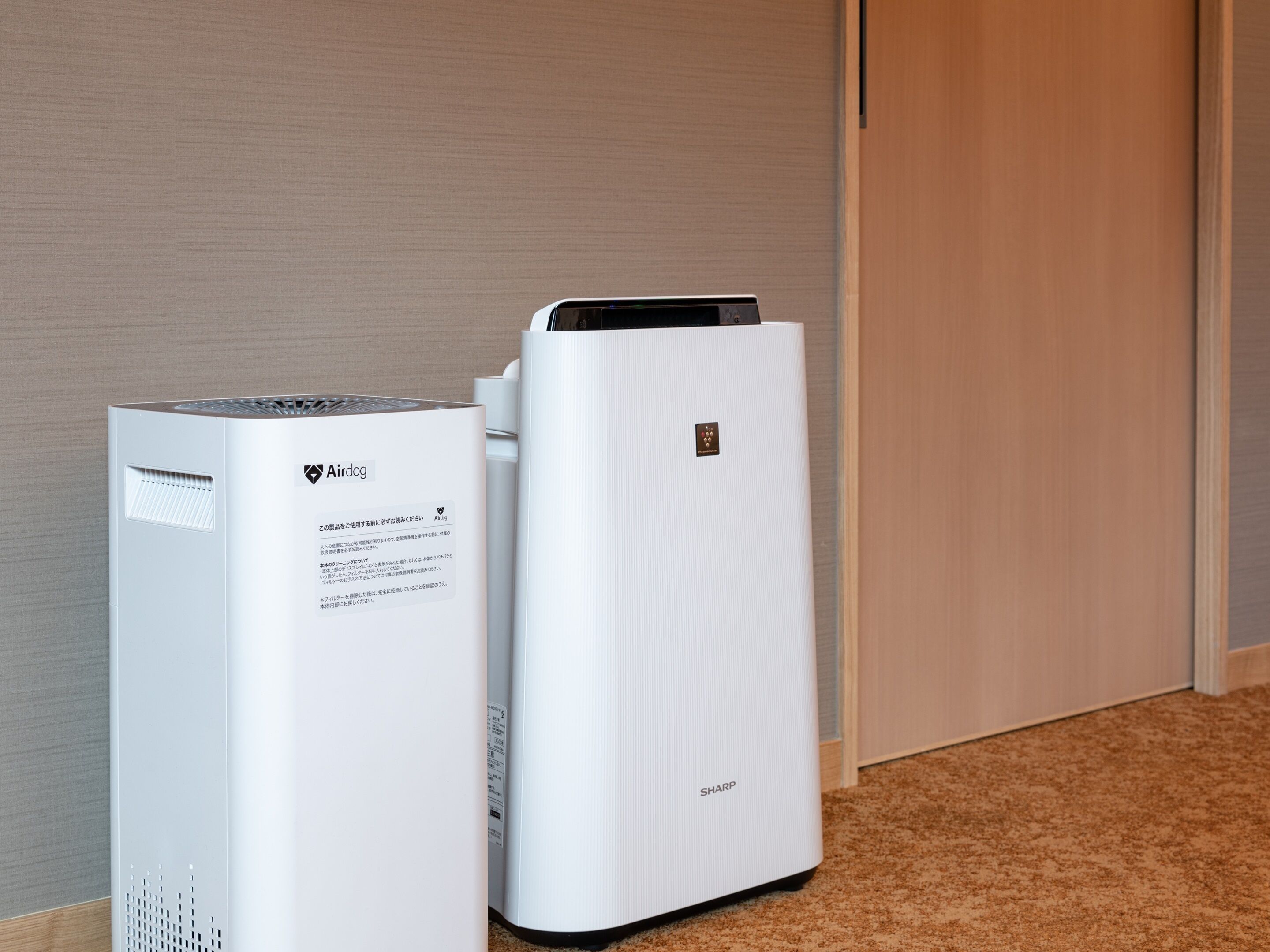 Two white air conditioning units with the brand Airdog and Sharp labels, placed against a beige wall and wooden door.