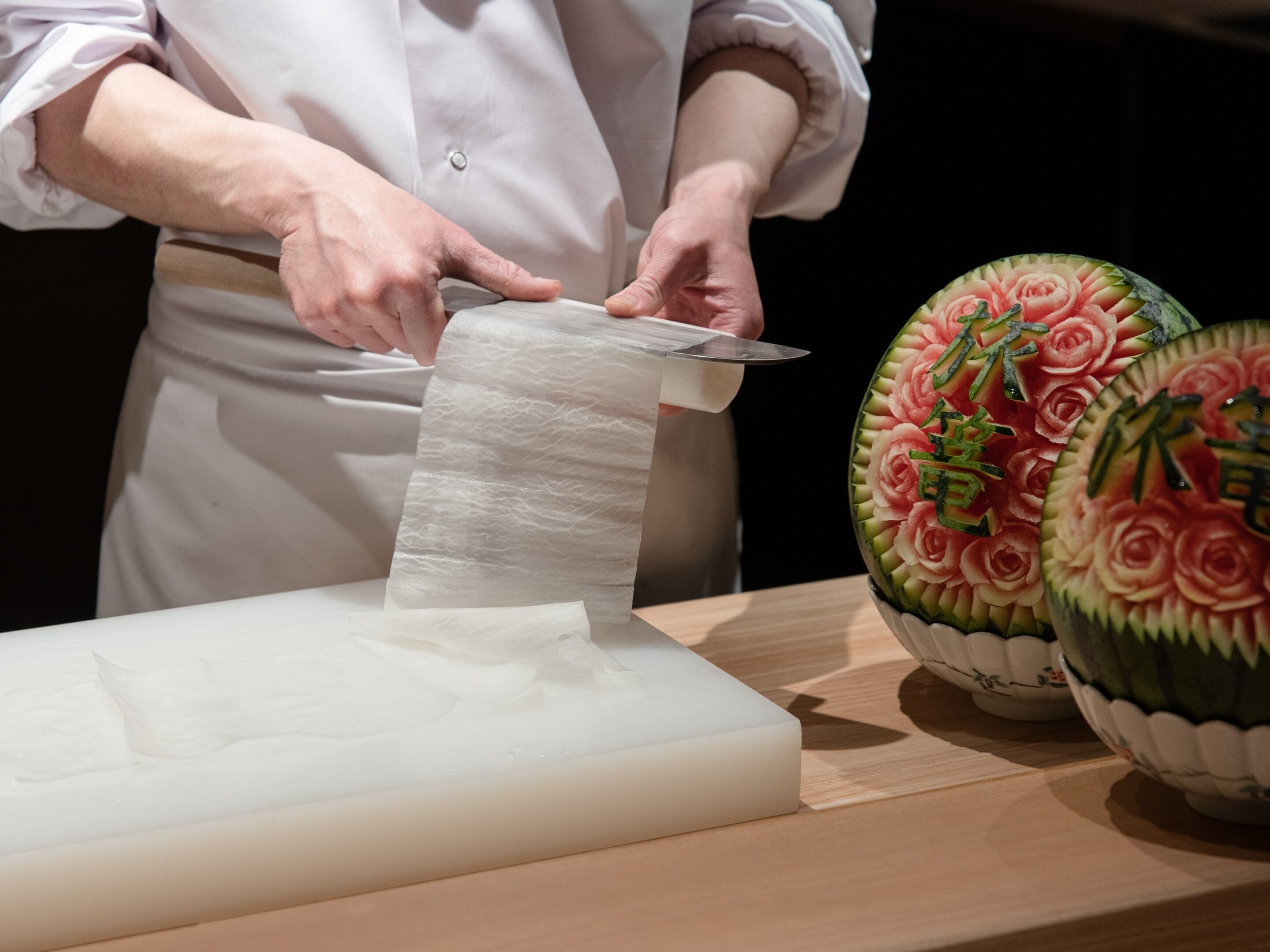 A person in a white chef's coat using a knife to cut a large white sheet of material on a wooden cutting board. There are two intricately carved watermelons with floral designs on the table beside the cutting board.