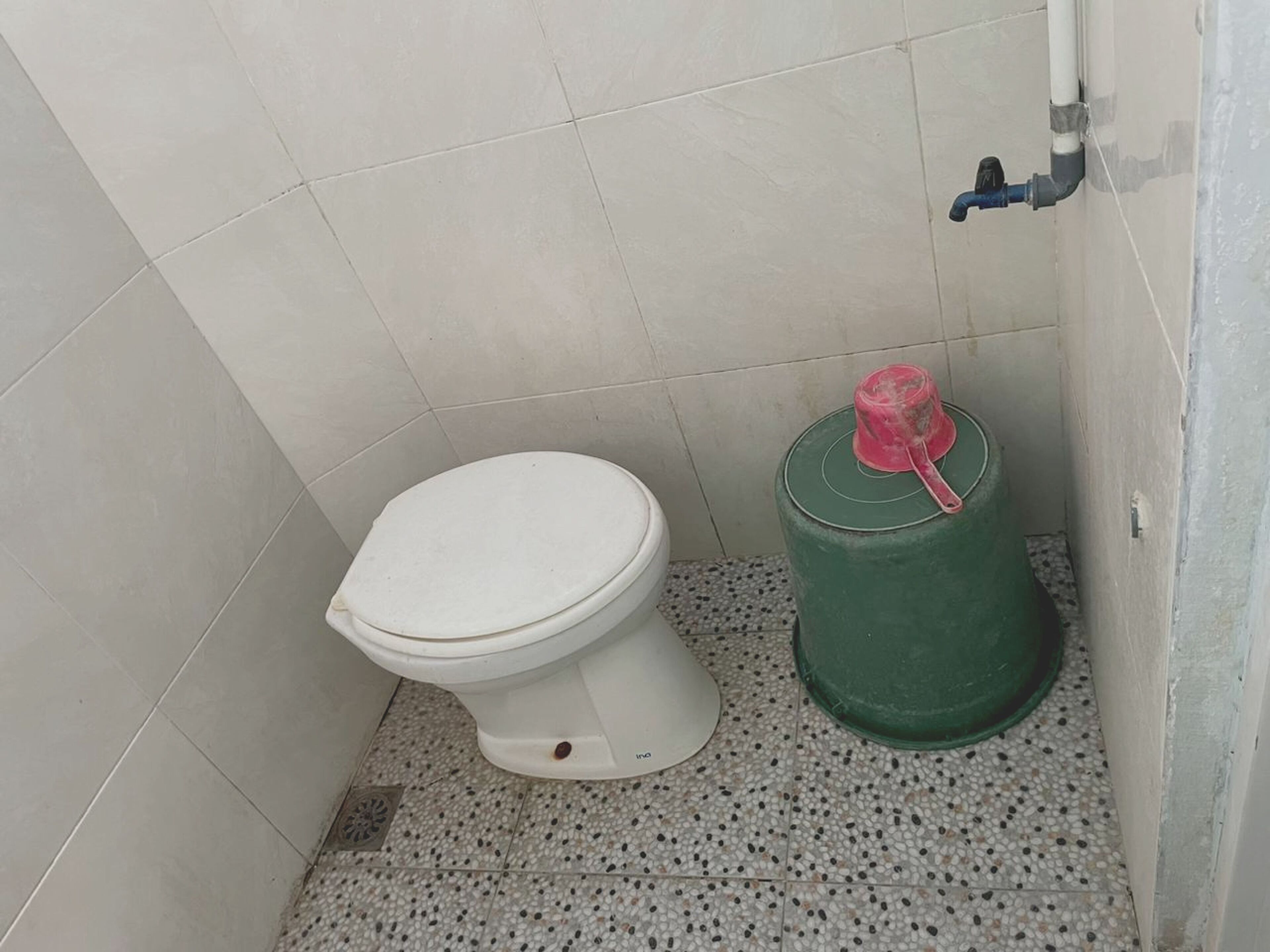 A small, tiled bathroom with a white toilet, a green water container, and a blue pipe.