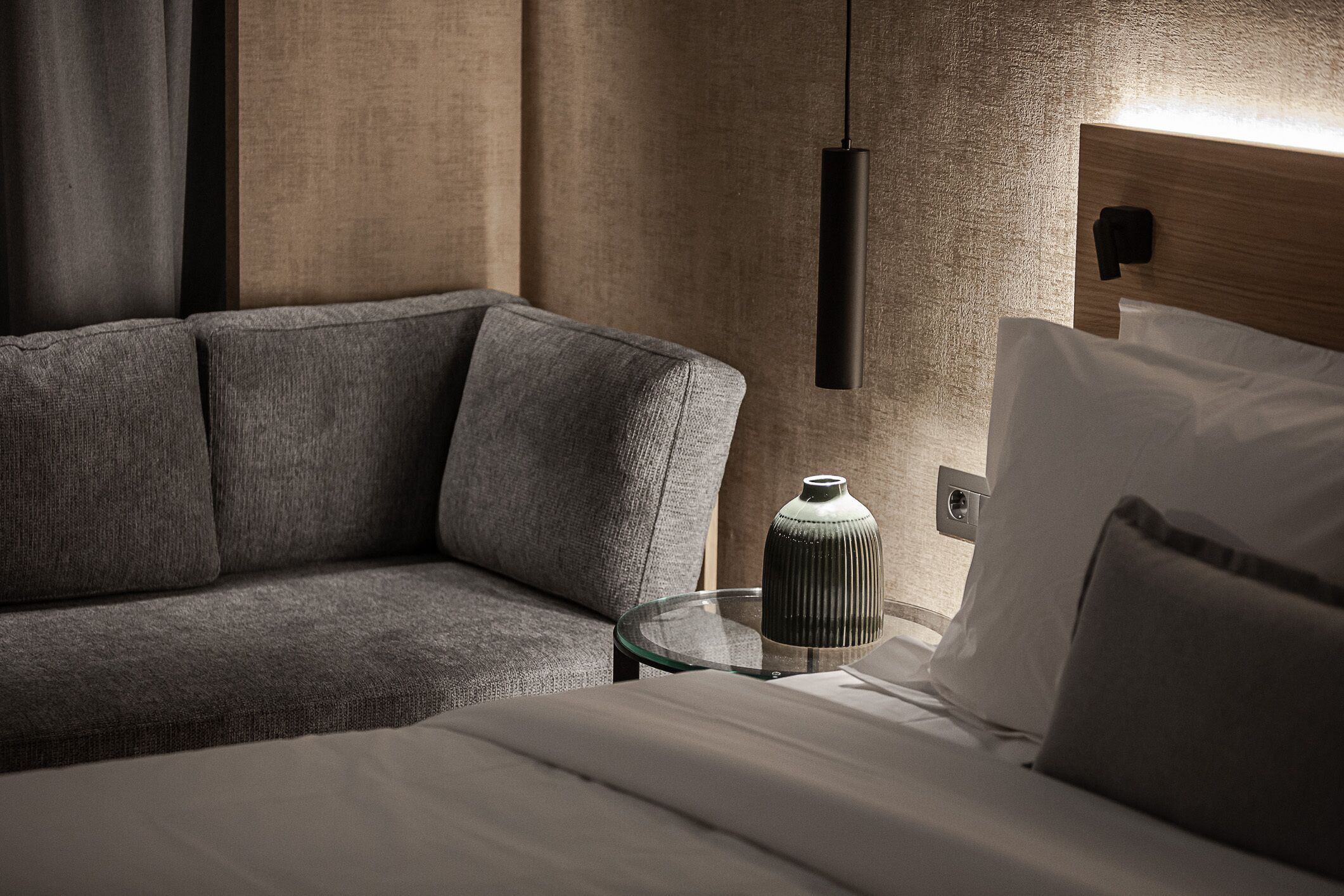 A modern hotel room with a sectional sofa, a glass side table, a decorative vase, and a wall-mounted light fixture.