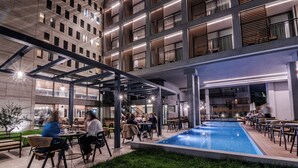 An indoor pool area with seating and tables, surrounded by a building with multiple floors and balconies. - Magnes Hotel (Volos)