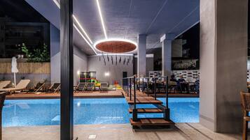 Un área de piscina interior moderna con sillones de descanso, un comedor y un diseño de techo único.