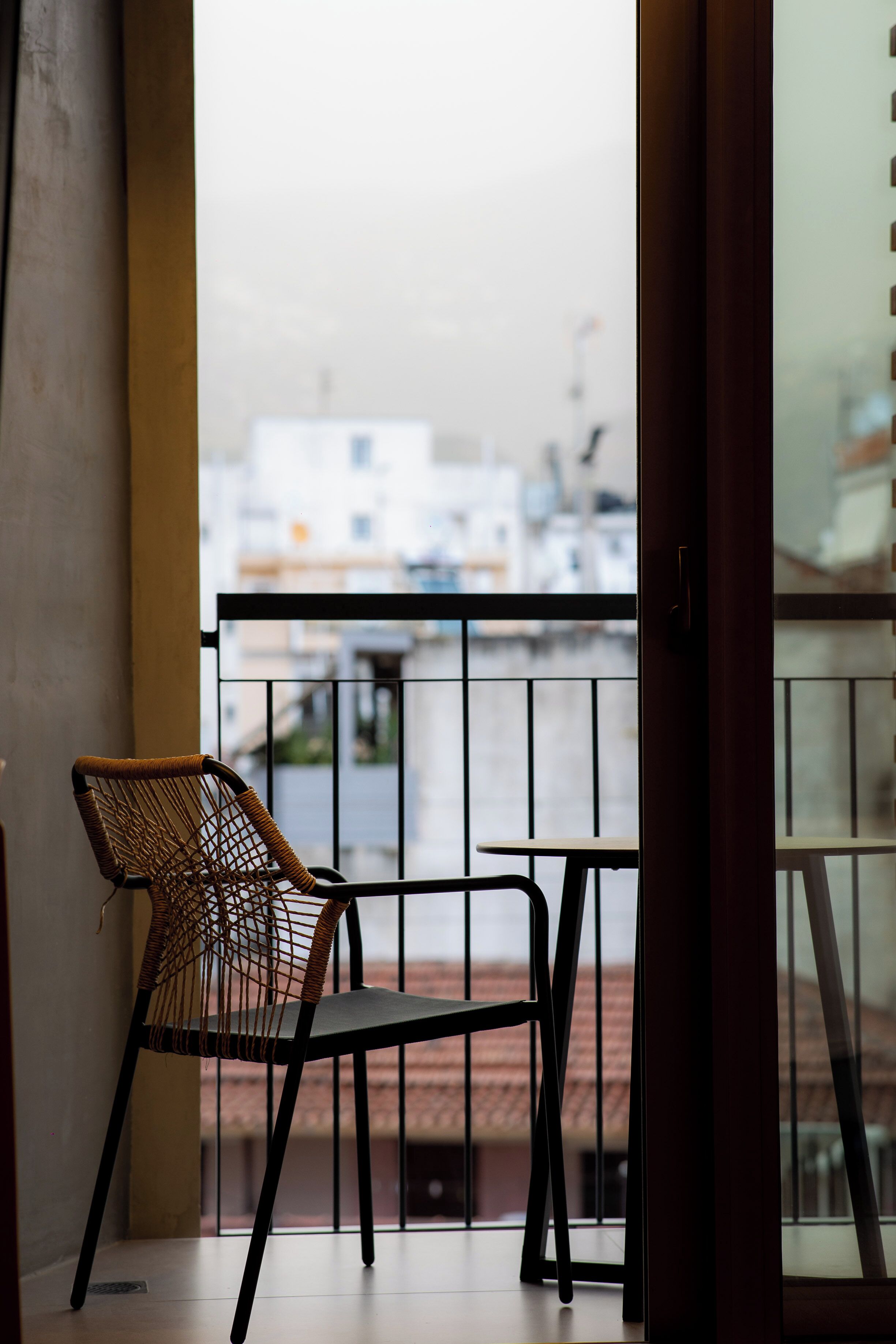 Un balcón con una silla de mimbre y una mesita pequeña, con vistas a un paisaje urbano con edificios y cielo despejado.