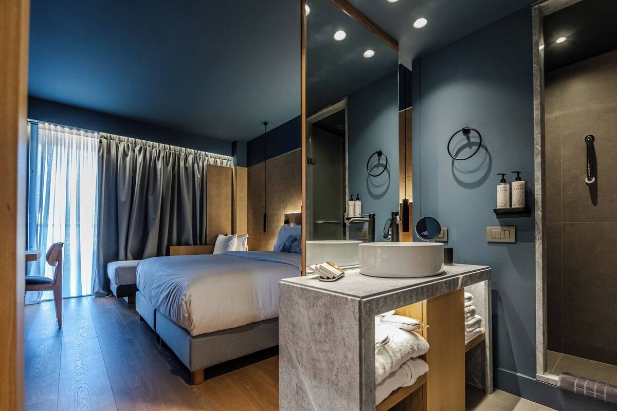 Habitación de hotel moderna con una cama grande, baño con lavamanos y espejo, y vista al exterior por la ventana.