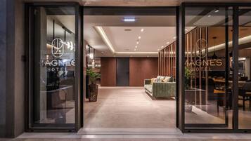 Entrada de hotel con puertas de cristal, zona de recepción y espacio de descanso con sofá y sillones.