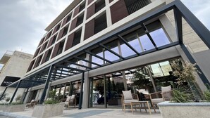A modern hotel with a glass entrance, outdoor seating, and a visible car in the background. - Magnes Hotel (Volos)