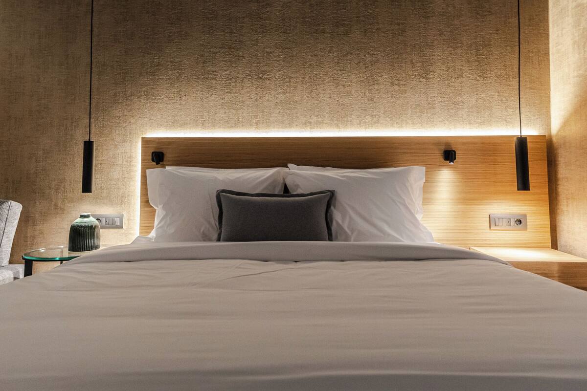 Una cama bien tendida con sábanas blancas y un cabecero de madera. Dos lámparas colgantes negras están sobre la cama.
