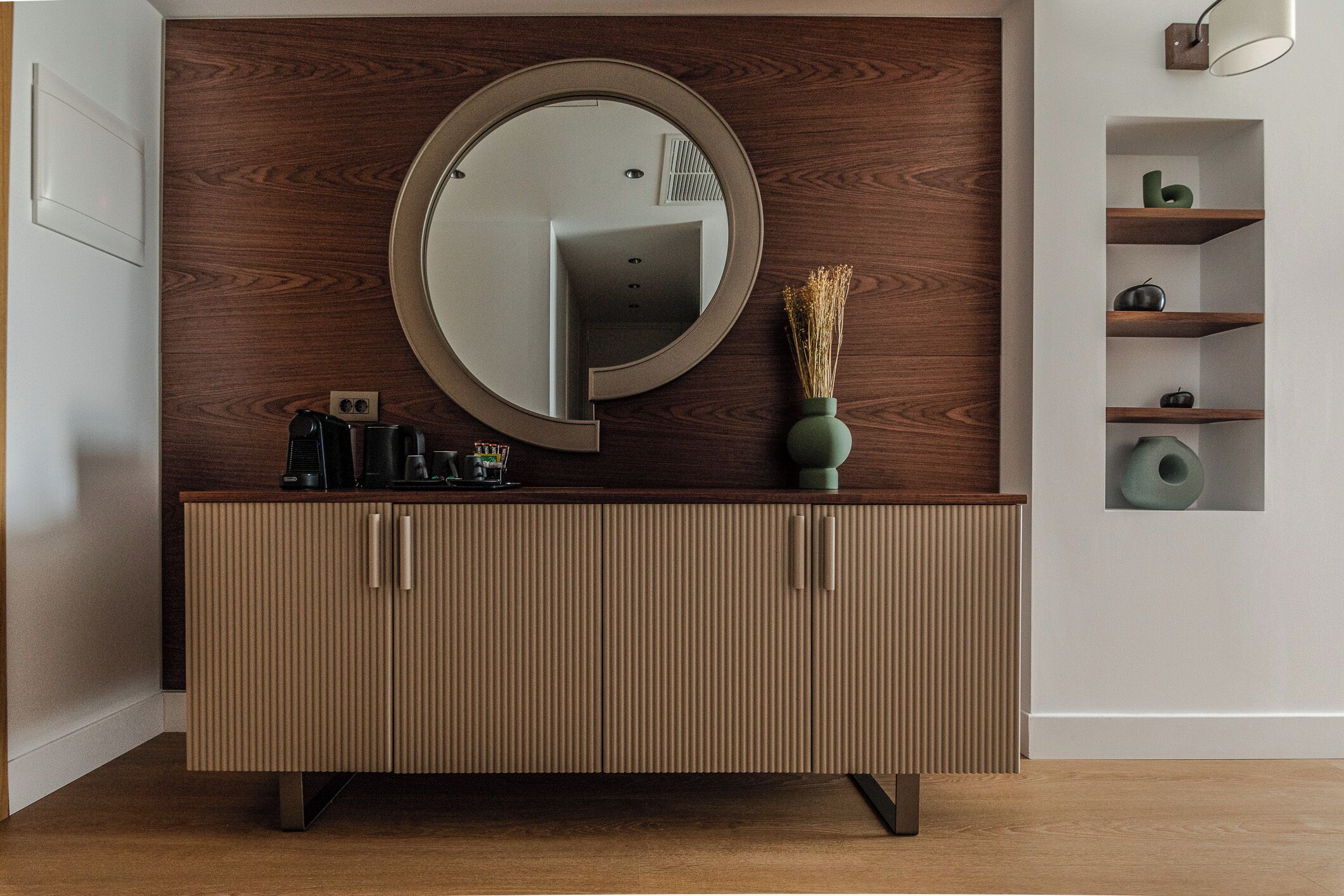A modern bathroom with a wooden vanity, a round mirror, and a built-in shelf with decorative items.