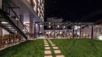 An outdoor seating area with tables and chairs, a grassy lawn, and a building with multiple floors and balconies.