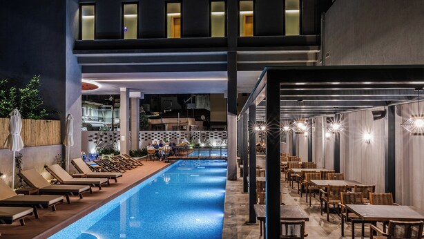 A swimming pool with lounge chairs and tables, surrounded by a covered outdoor area with a view of the cityscape. - Magnes Hotel (Volos)