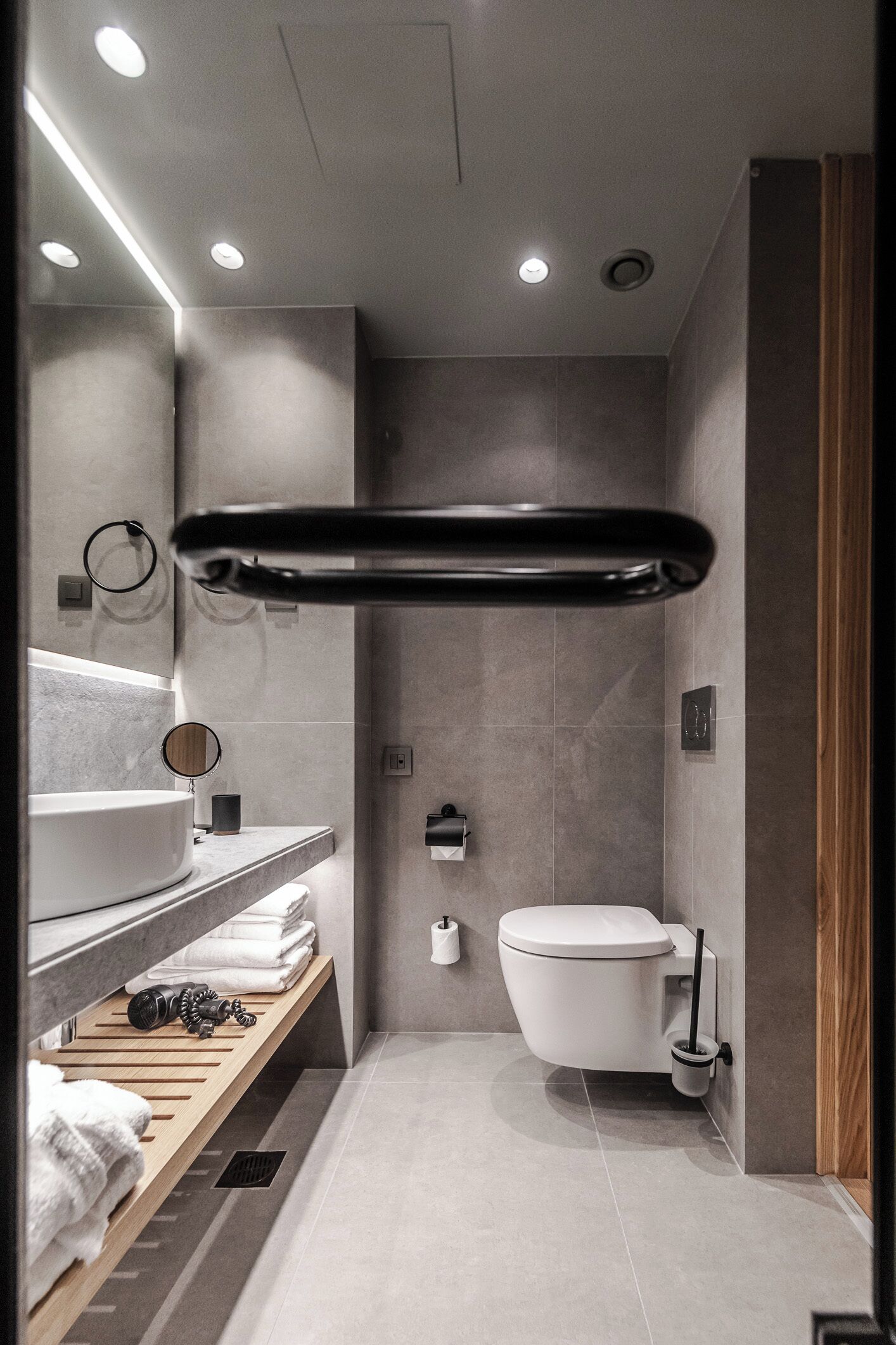 A modern bathroom with a white sink, mirror, and toilet. There is a towel rack above the sink and a shelf with towels and toiletries.