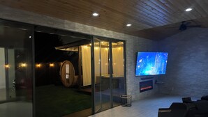 Un interior moderno con entrada por puerta de vidrio, techo de madera, televisor montado y una pequeña zona de estar.