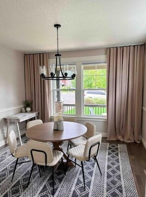 A dining area with a round table, six chairs, a chandelier, a window with curtains, and a view of greenery outside. - Mt Hood Luxury Home with Hot tub Game room & More (Gresham)
