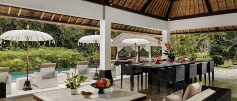 Un área de comedor al aire libre con mobiliario de mimbre, sombrillas y una piscina al fondo.