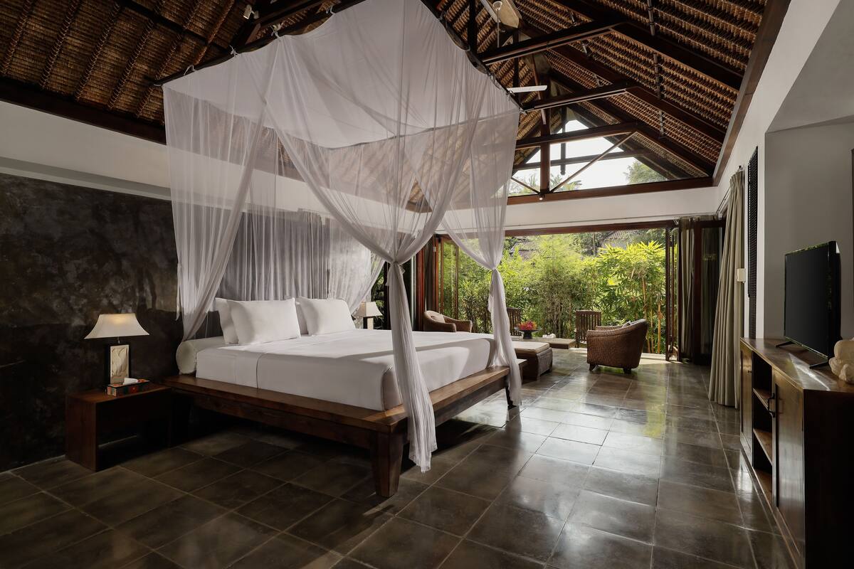 A spacious bedroom with a large bed, a nightstand, a TV, and a view of greenery outside.