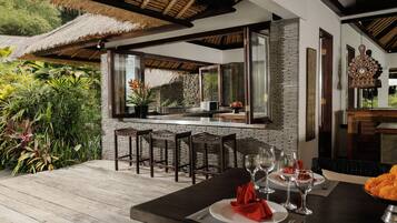 An outdoor dining area with a thatched roof, stone walls, and a view of lush greenery.