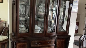 Un mueble de madera oscura con puertas de cristal y cajones, situado en una habitación con suelo de baldosas y una mesa de comedor.