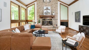 A spacious living room with a stone fireplace, large windows, and a high ceiling with exposed beams.