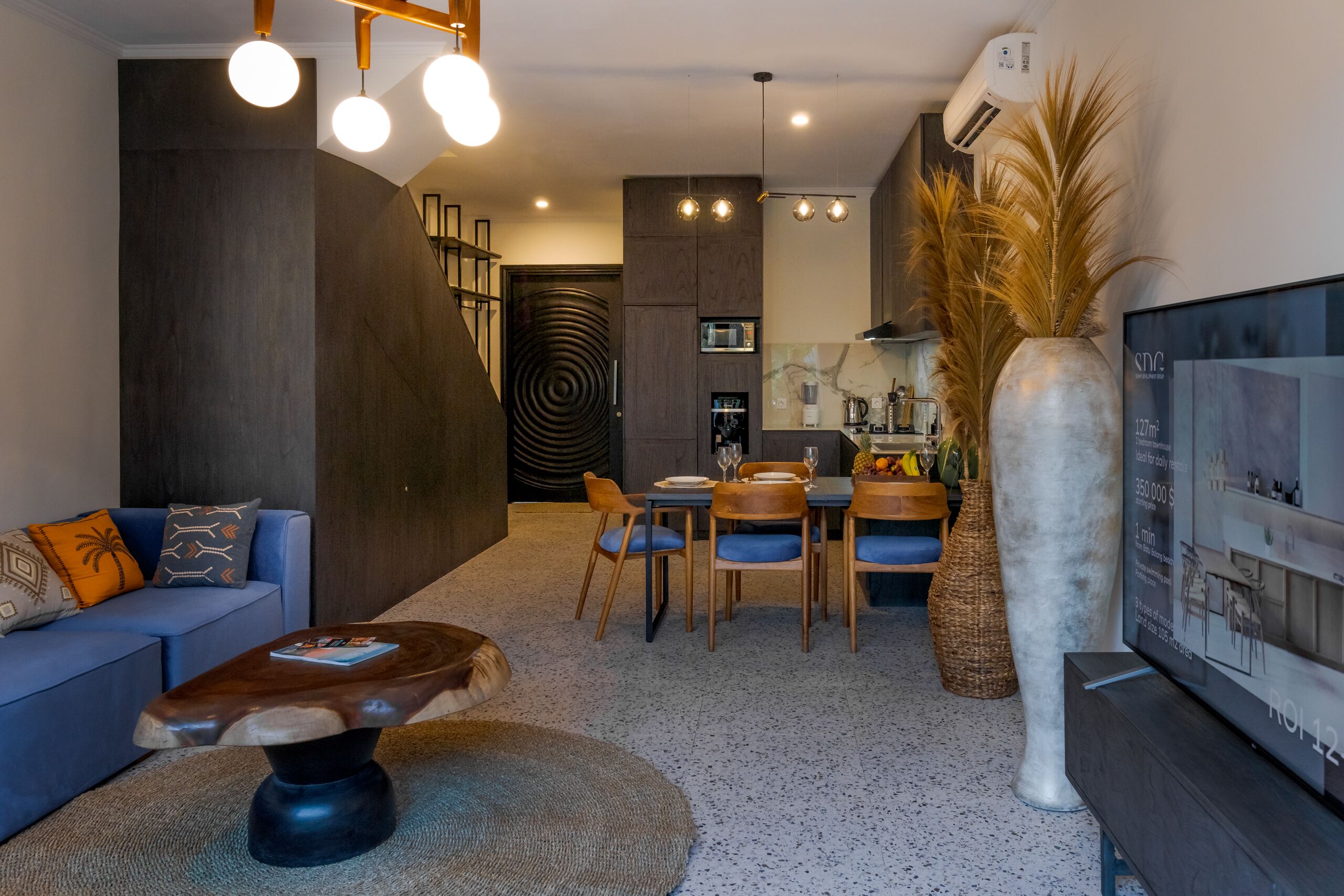 A modern living room with a dark wood accent wall, a blue sofa, a coffee table, and a dining area with chairs and a table.
