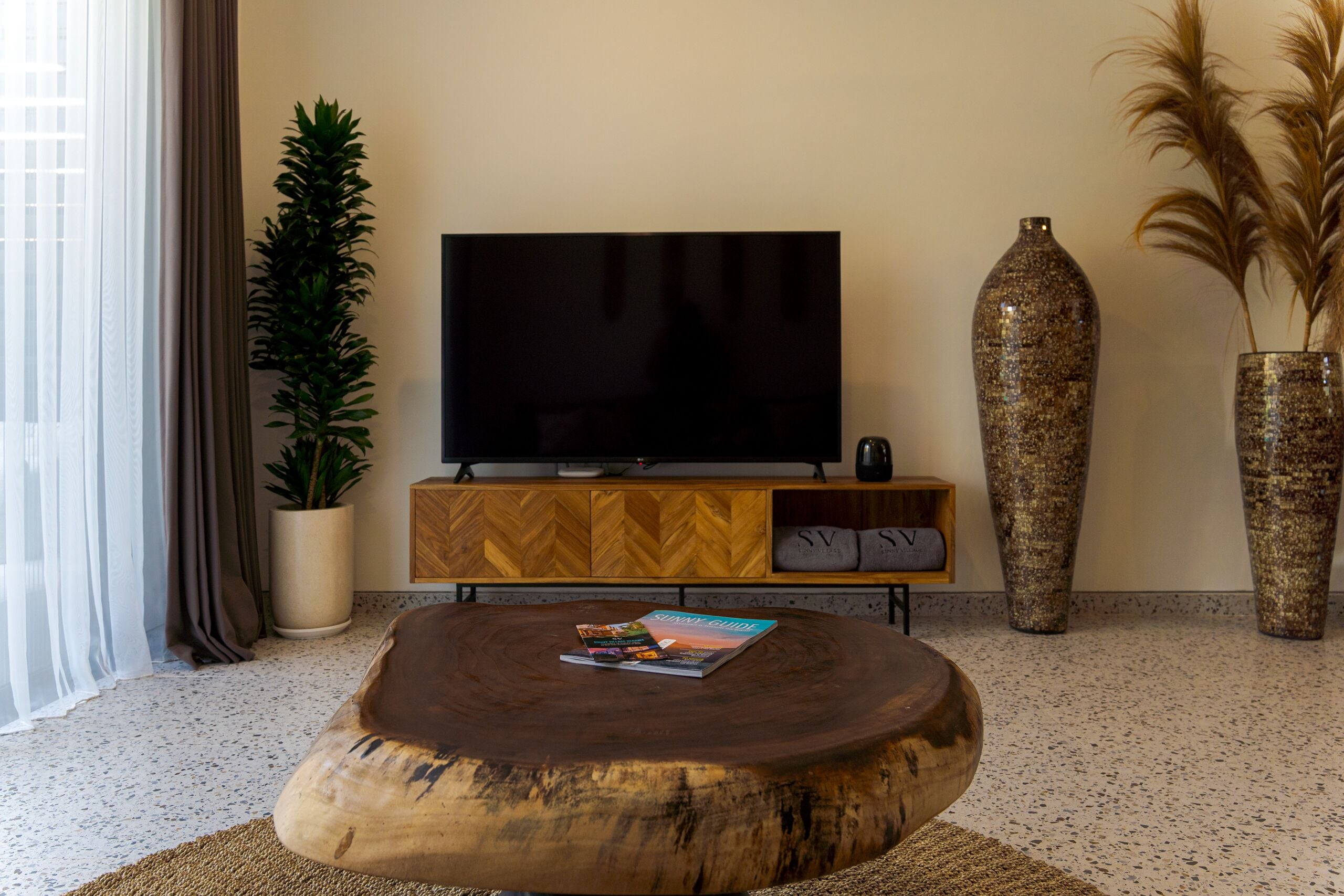 Una sala de estar moderna con una mesa redonda de madera, un televisor de pantalla plana sobre un mueble de madera, una planta grande en maceta y dos jarrones altos.