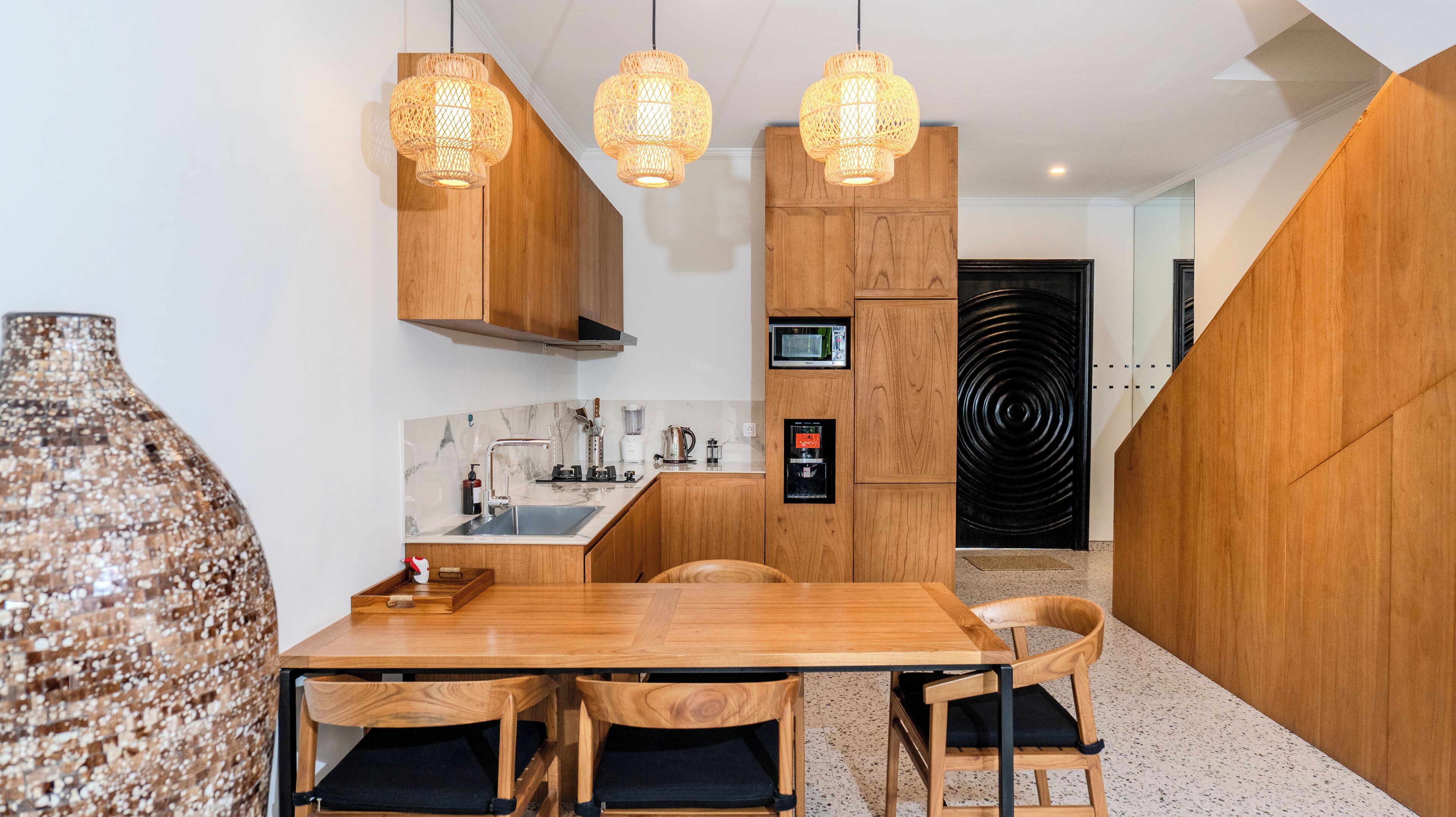 Una cocina moderna con armarios de madera, encimera de mármol y microondas integrado. Hay tres lámparas colgantes, un jarrón grande y una mesa de comedor con sillas.