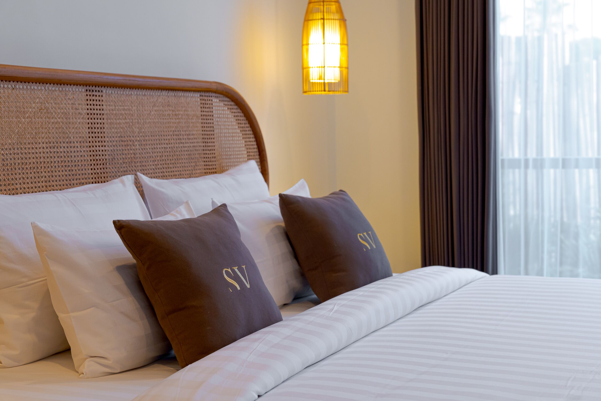 Una cama bien tendida con almohadas blancas y marrones, un cabecero tejido y una lámpara de pared.