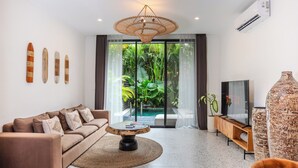 A modern living room with a sofa, coffee table, TV, and a large window overlooking a lush garden. - Sunny Village Batu Bolong (Canggu)