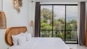 A bedroom with a large bed, a wicker headboard, and a view of greenery through a large window. - Sunny Village Batu Bolong (Canggu)