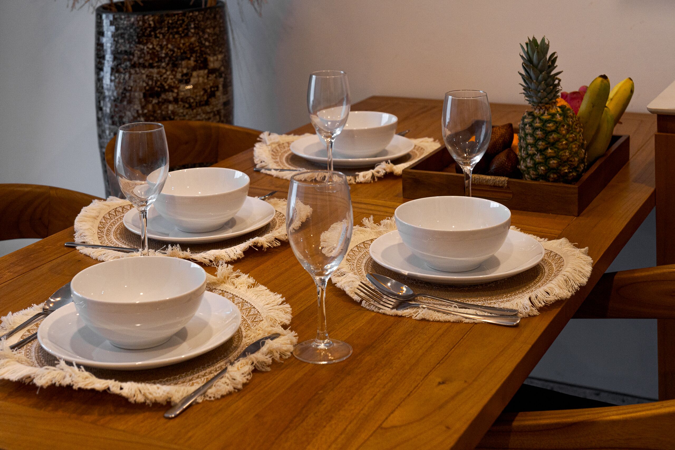 Une table à manger est dressée avec des bols, des assiettes et des verres à vin blancs, accompagnée d’un saladier rempli de bananes et d’un ananas.