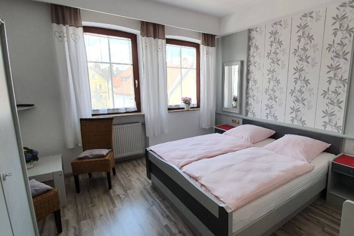 Un dormitorio con una cama doble, una mesita de noche, una silla, un ventanal con cortinas y un radiador.