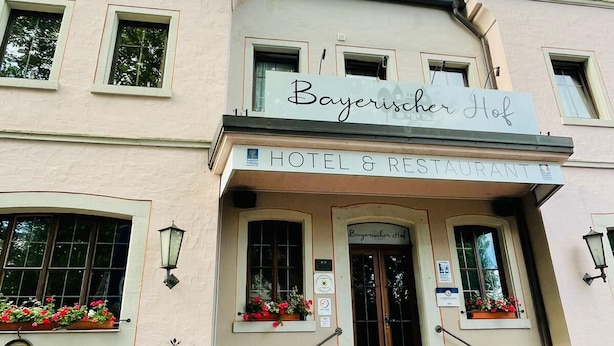 Un hôtel avec une enseigne indiquant « Bayerischer Hof » et une autre indiquant « HÔTEL ET RESTAURANT ». On y trouve des tables et des chaises à l’extérieur, ainsi que des jardinières sur les rebords des fenêtres.