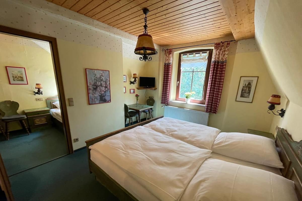 Un dormitorio con techo de madera, cama con ropa de cama blanca, escritorio con silla y ventana con cortinas.