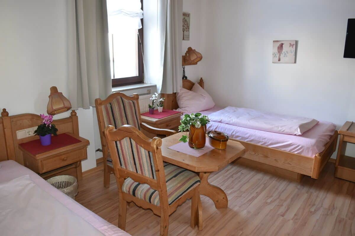 Un dormitorio acogedor con dos camas, una silla de madera, una mesita con un florero y una ventana con cortinas.