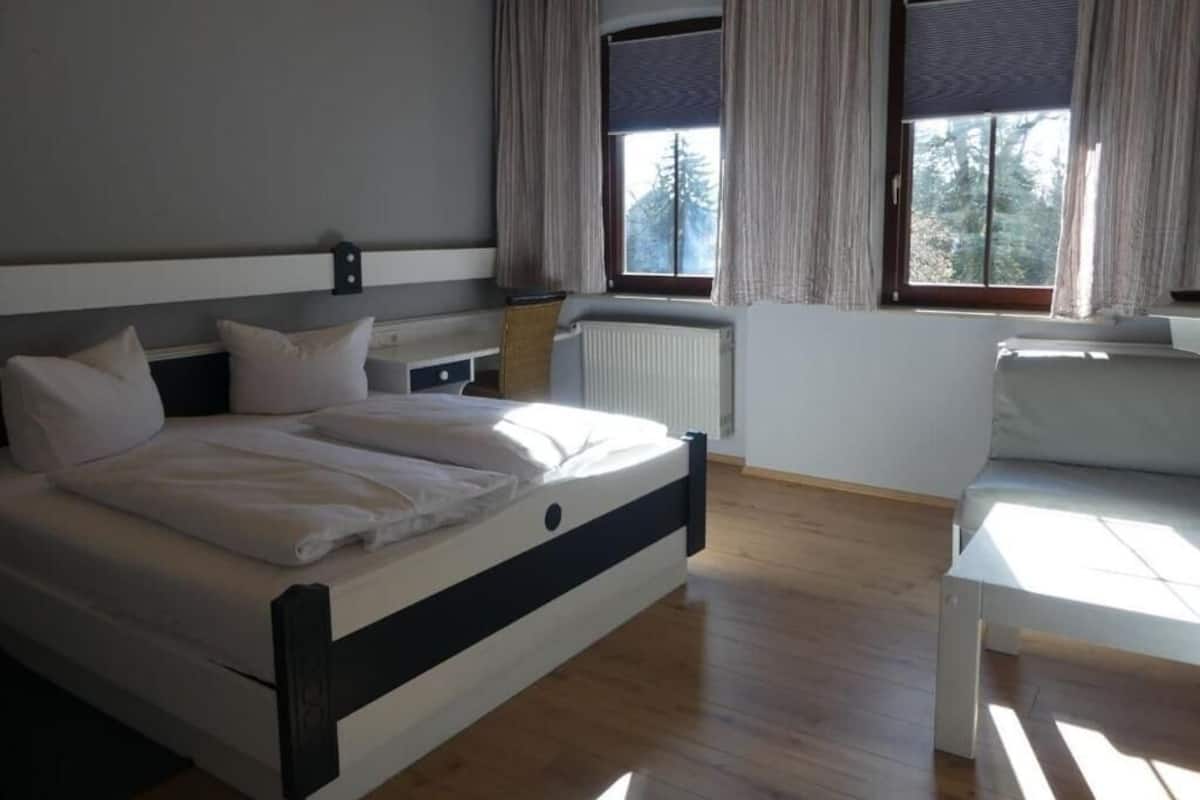 Un dormitorio moderno con una cama grande, dos ventanas con cortinas, un radiador y una mesita.