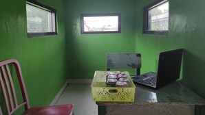 A room with green walls, two windows, a desk with a laptop, a chair, and a yellow crate with jars. - OYO 90333 Wisma Tenang Jaya Syariah (Jatiwangi)